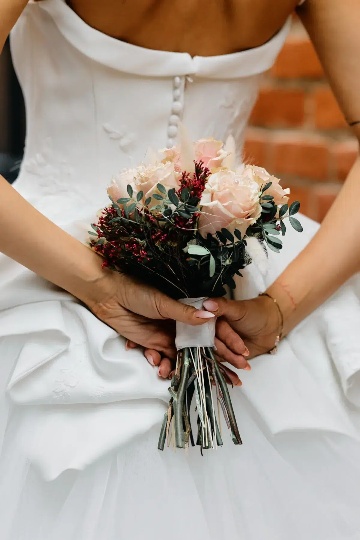 Close detail of a bridal gown's button back and blush bouquet held behind the bride, photographed against brick in Luxembourg.