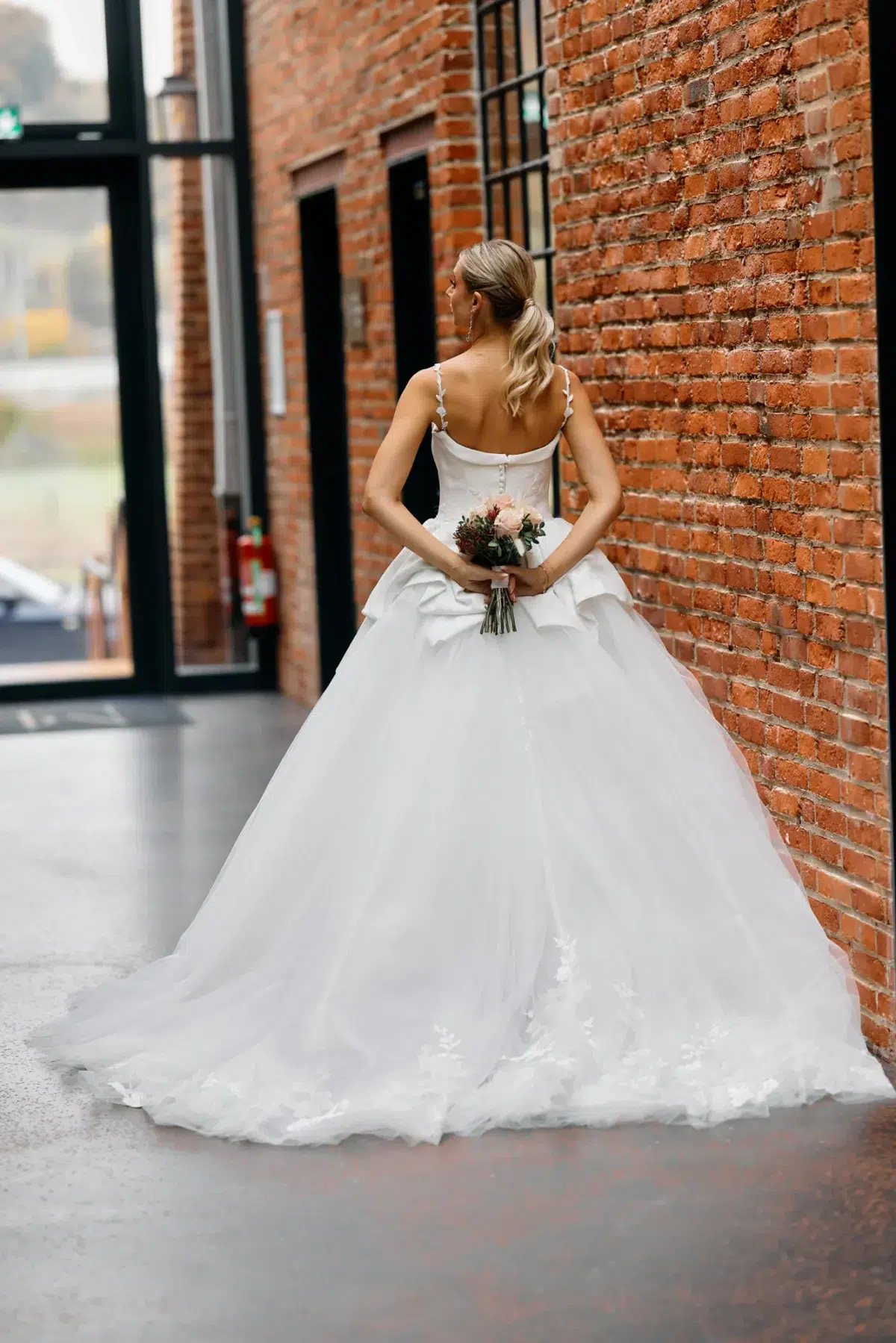 Back view of a bride in a bow-strap ballgown holding her bouquet behind her back against an exposed brick wall, Luxembourg wedding photography.