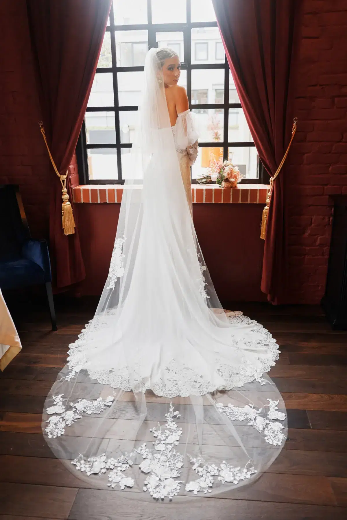 Bride standing at an arched window wearing a lace-edged cathedral veil, bridal portrait in Luxembourg.