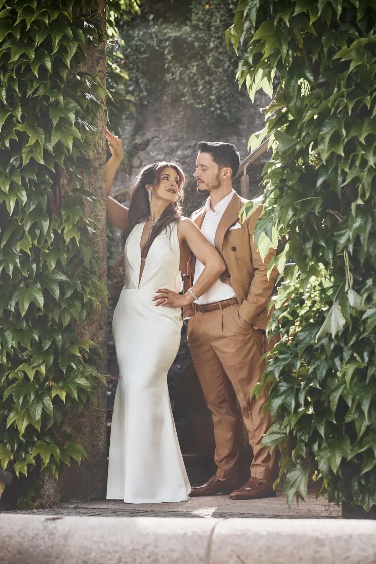 Bride and groom share a playful, confident pose between two ivy-covered columns, a striking portrait from their wedding day.