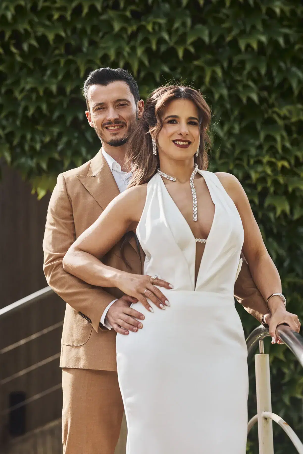 Bride and groom pose together in front of a lush ivy wall, showcasing the bride's diamond necklace and gown during their wedding portraits.