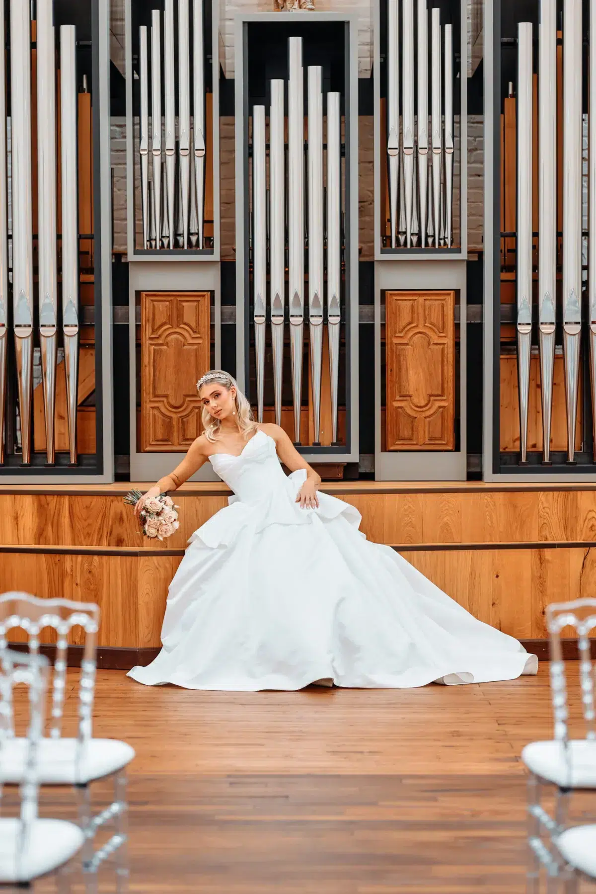 Bride in a sweetheart ballgown sitting on the altar steps in front of a pipe organ, holding a blush rose bouquet, bridal portrait in Luxembourg.