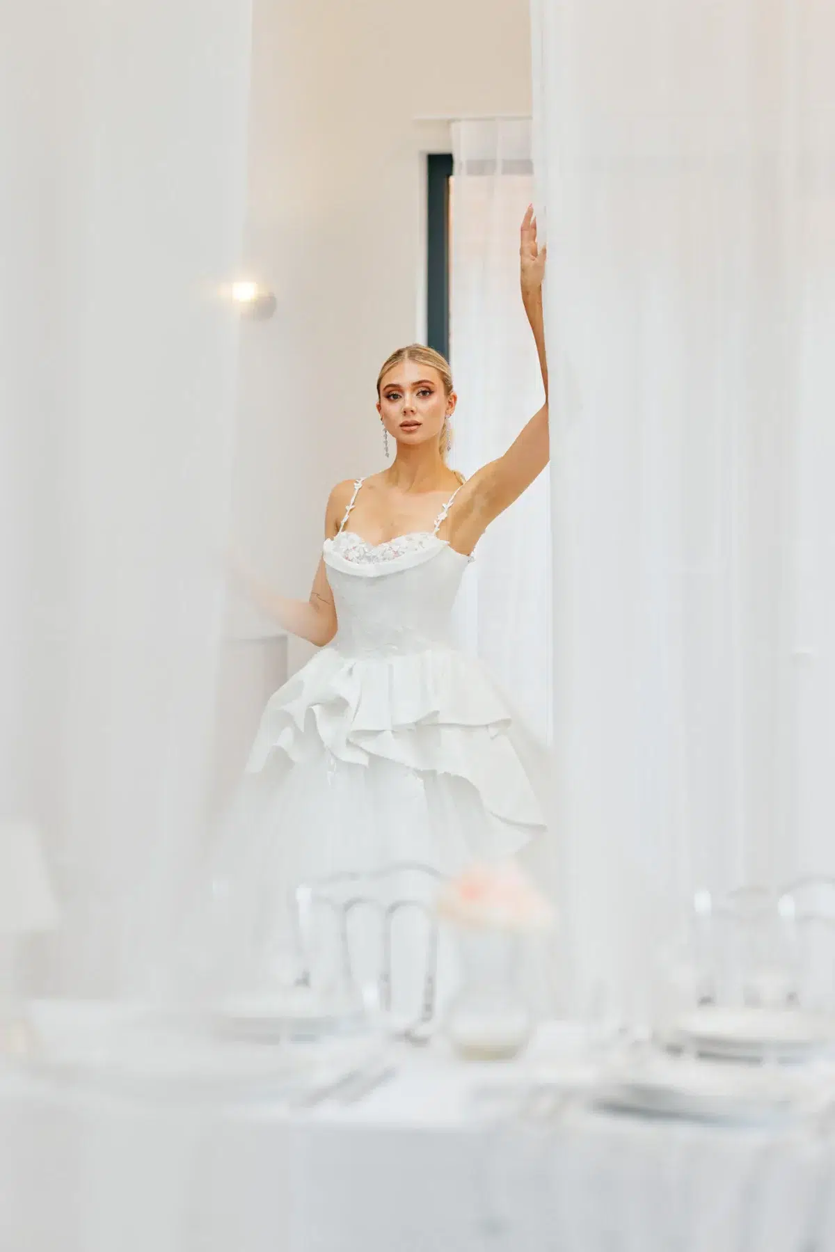 Full-length bridal portrait with the bride reaching for a sheer curtain in a bright white interior, Luxembourg wedding photography.