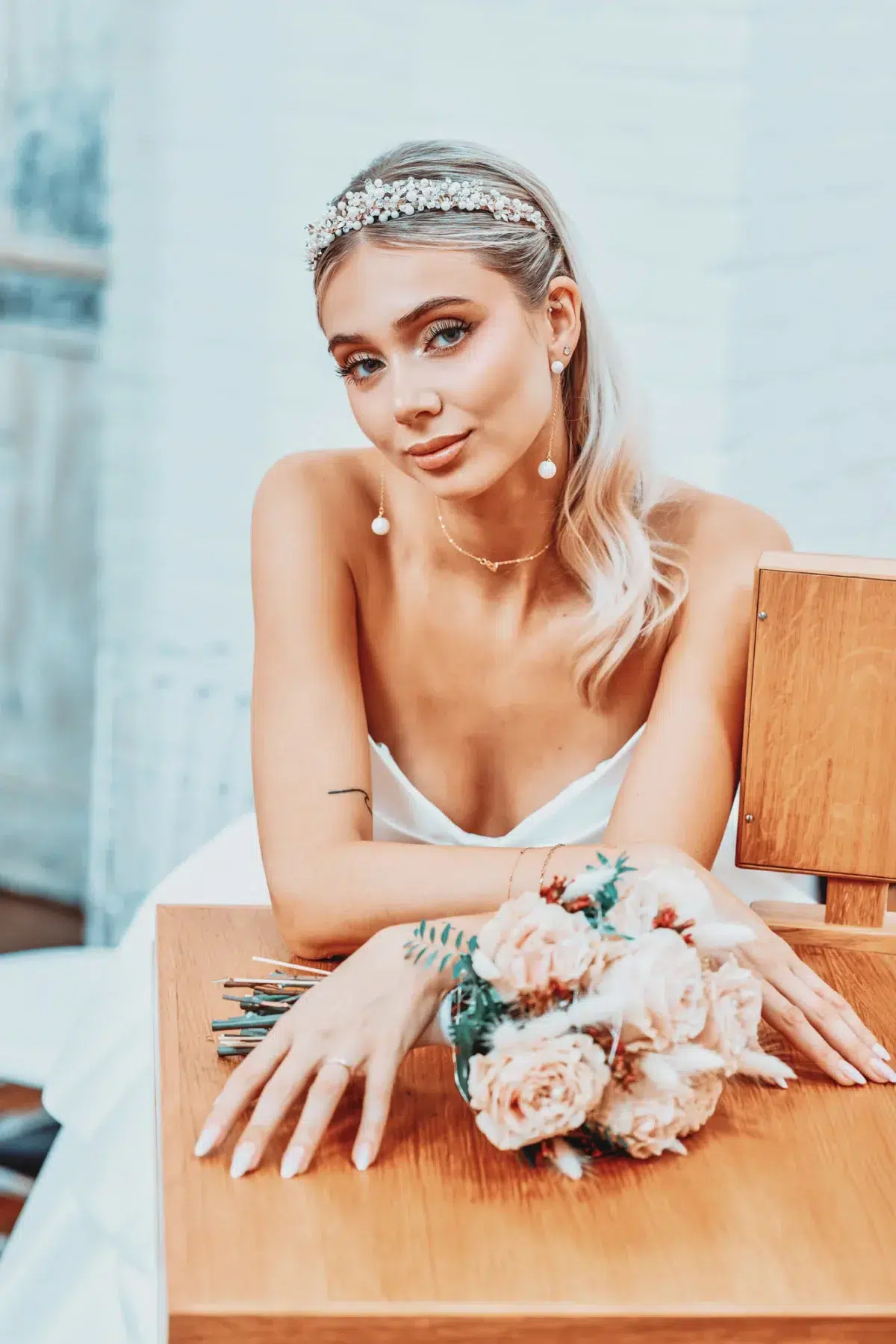 Close bridal portrait with pearl drop earrings and a blush bouquet resting on a wooden table, Luxembourg wedding photography.