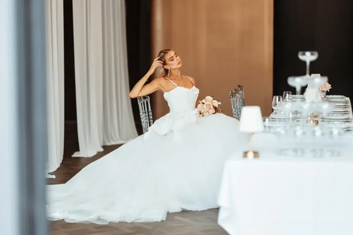 Bride seated at an elegant reception table with a tan curtain backdrop and crystal chairs, running her hand through her hair, Luxembourg wedding photography.