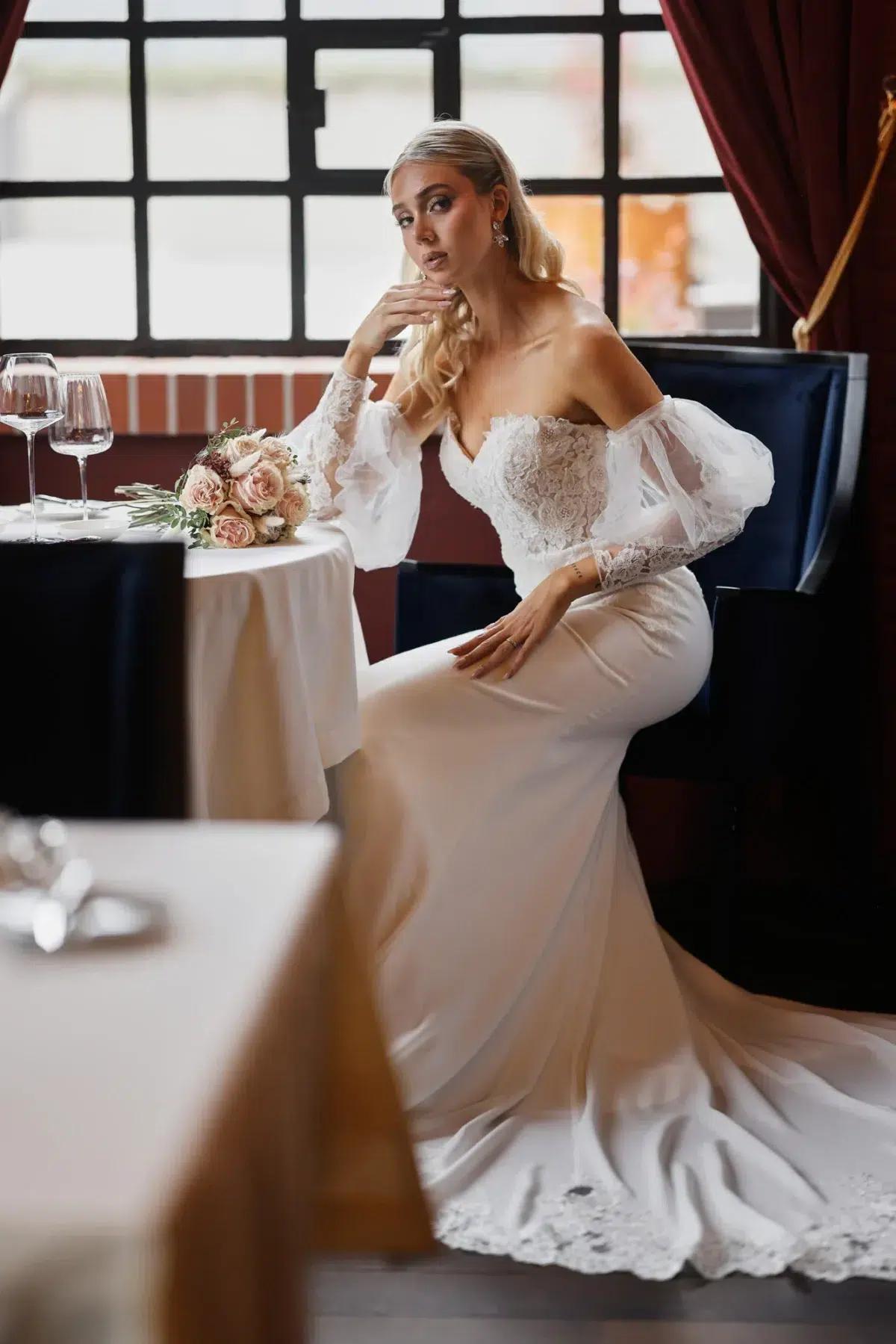 Bride in an off-shoulder lace gown with sheer puff sleeves seated at an elegant restaurant table with red velvet curtains, Luxembourg wedding photography.